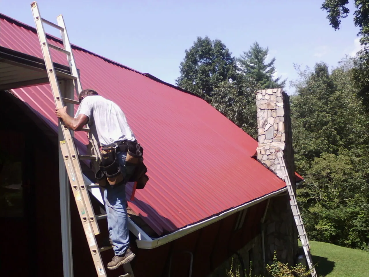 Roofer on a ladder beside a red metal roof and stone chimney — Soffit & Fascia Repair by Roofing Brothers in Tiverton
